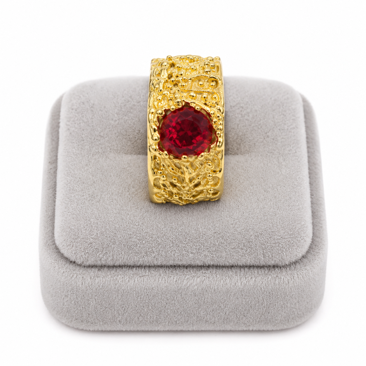 Anillo dorado ancho con relieve ornamental y piedra roja central.